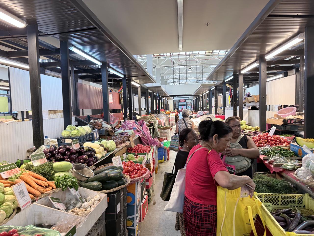 Shoppers at the vegetable market in the Victoria Urban Terminal, stalls piled with cabbages, eggplants, tomatoes and carrots, each with a handwritten price label